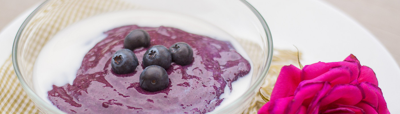 Pöytään on katettu lautanen, jolla on punainen ruusu ja lasikipossa jogurttia ja mustikoita.