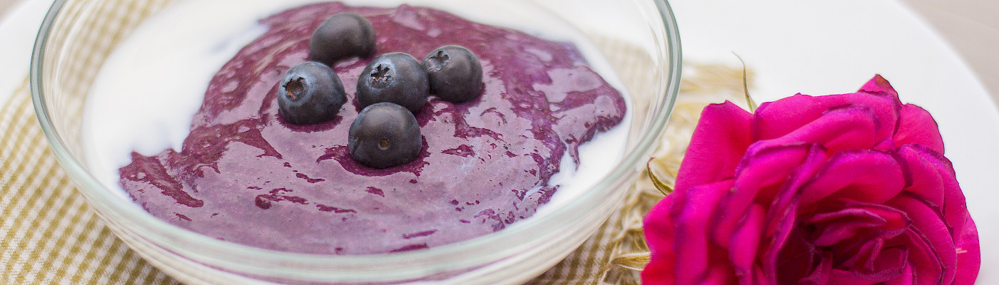 Pöytään on katettu lautanen, jolla on punainen ruusu ja lasikipossa jogurttia ja mustikoita.